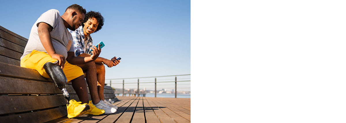 Two male friends sitting on a bench looking at their phones, one man has a prosthetic leg