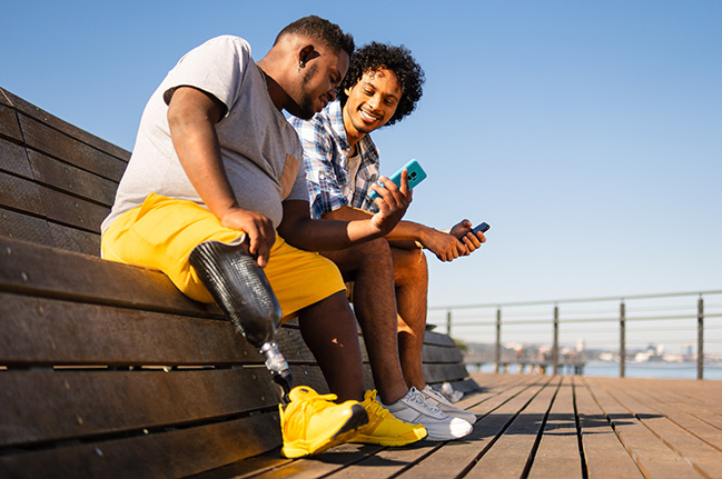 Two male friends sitting on a bench looking at their phones, one man has a prosthetic leg