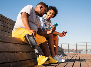 Two friends looking at phone outside on a bench, one has prosthetic leg